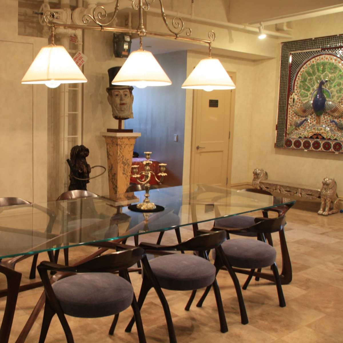 A modern dining room features a large glass table surrounded by eight dark wooden chairs, with a sculpture and decorative wall art in the background.