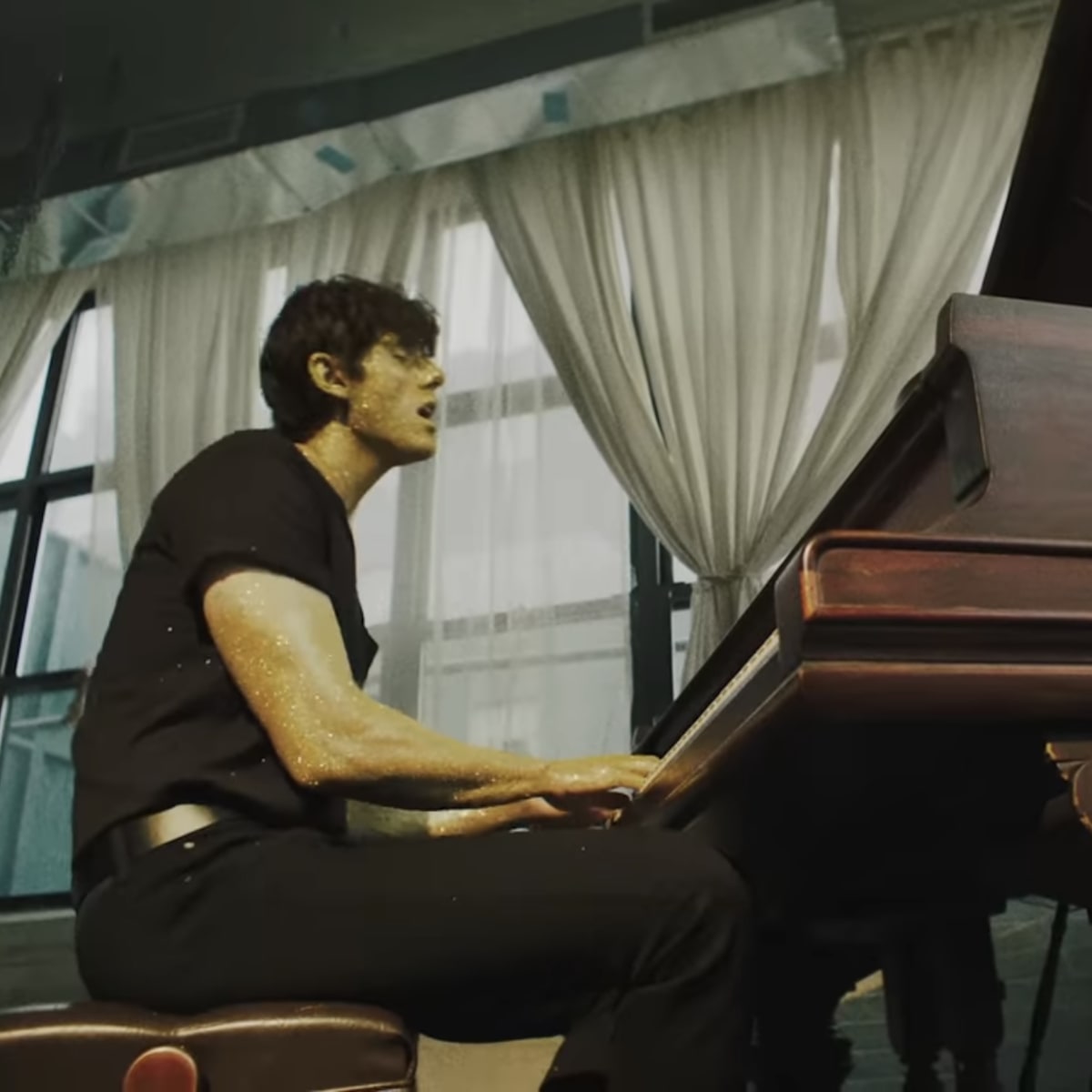 A person plays a grand piano in a room with large windows and white curtains.