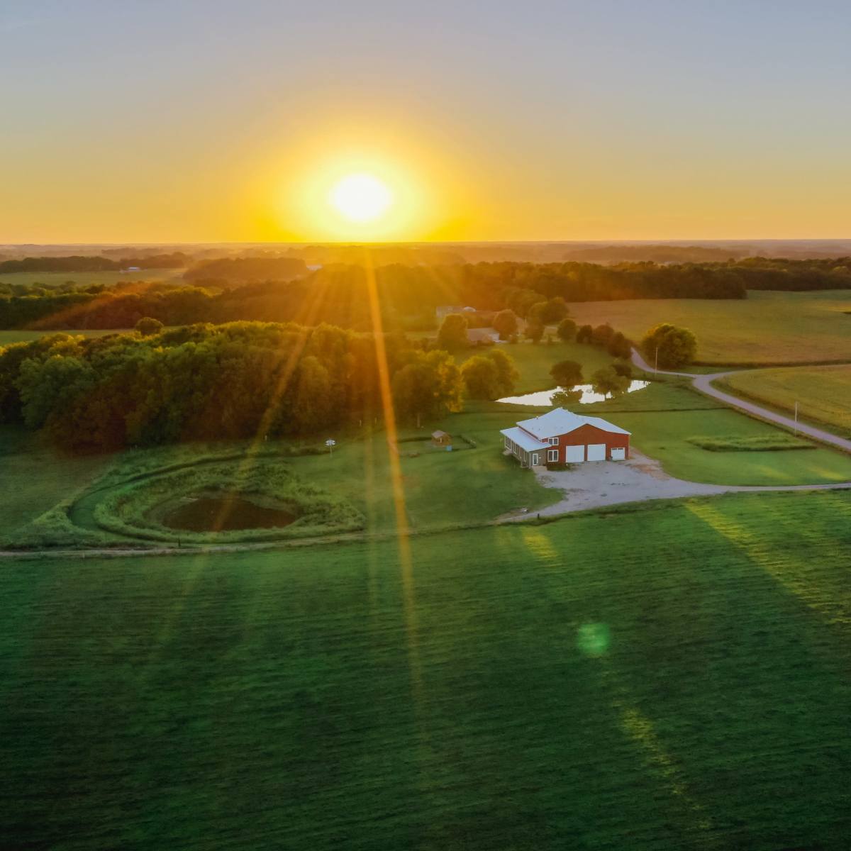 A farmhouse is situated in a lush green field with a pond, illuminated by the rising sun in the background.