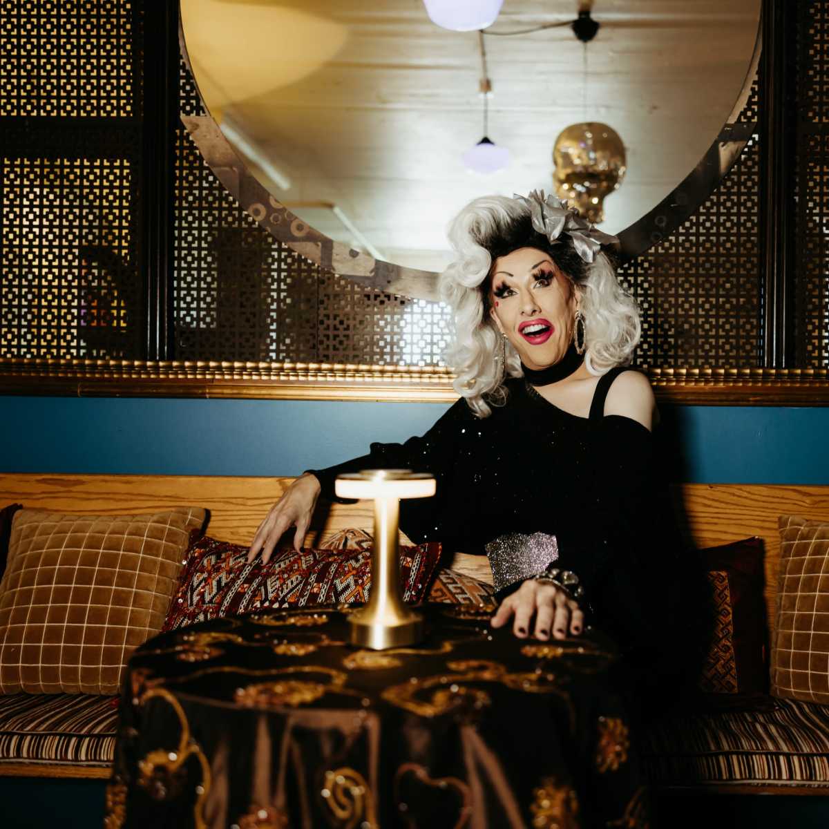A person with white hair in a black outfit sits at a table with a lamp, surrounded by patterned cushions in a dimly lit room.