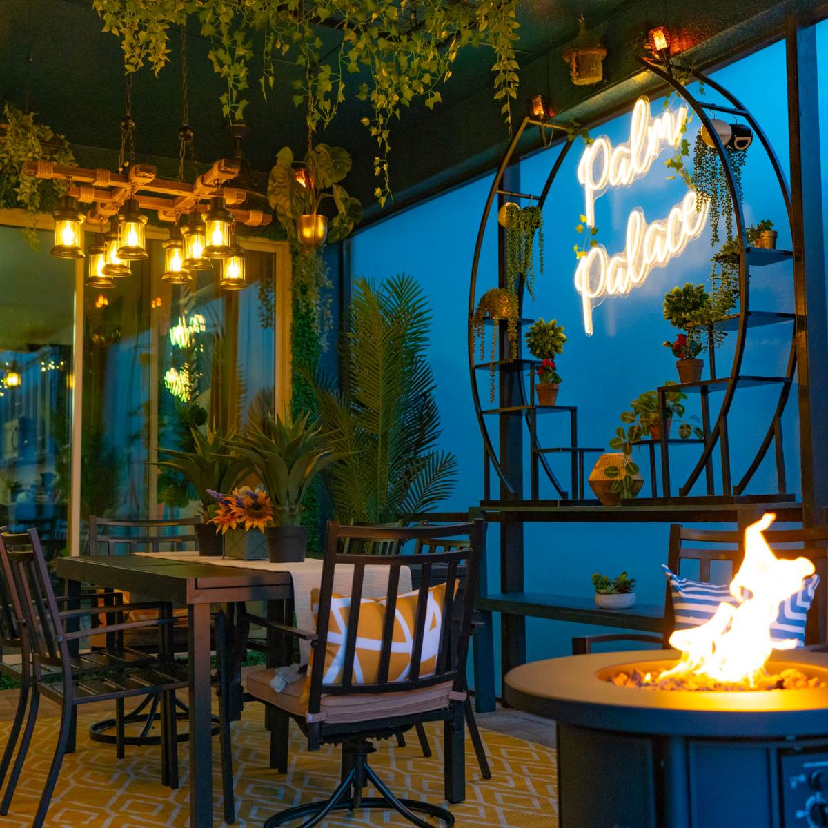 A cozy outdoor dining area featuring black tables and chairs, decorative plants, and a neon sign that reads "Palace."