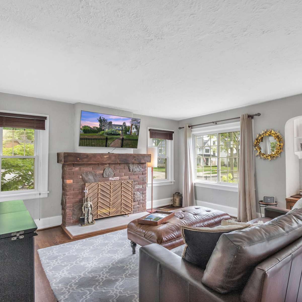 A living room features a brown leather sofa, a stone fireplace, and large windows with greenery visible outside.