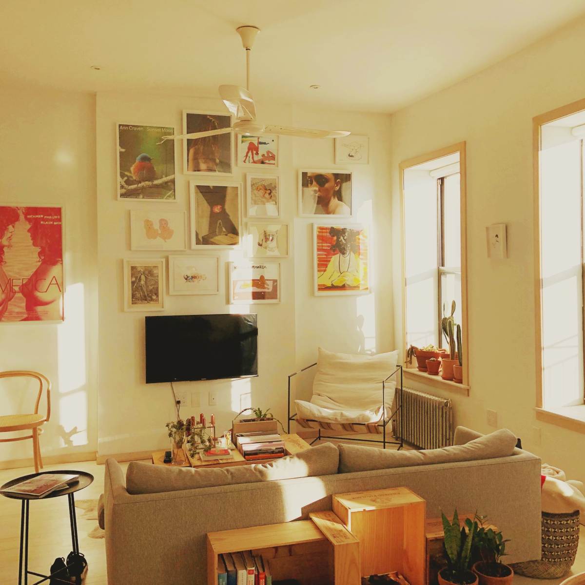 A brightly lit living room with a sofa, a wall adorned with various framed artworks, and large windows letting in natural light.