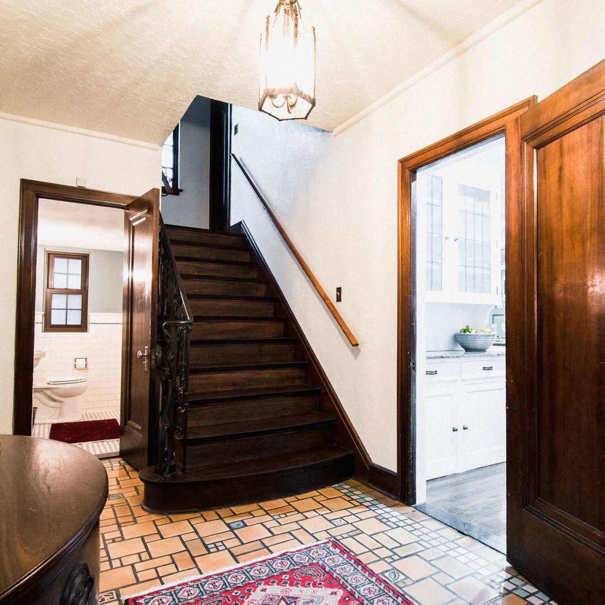 An entryway with a wooden staircase leading to an upper floor, a decorative rug on the tiled floor, and doorways leading to a bathroom and kitchen.