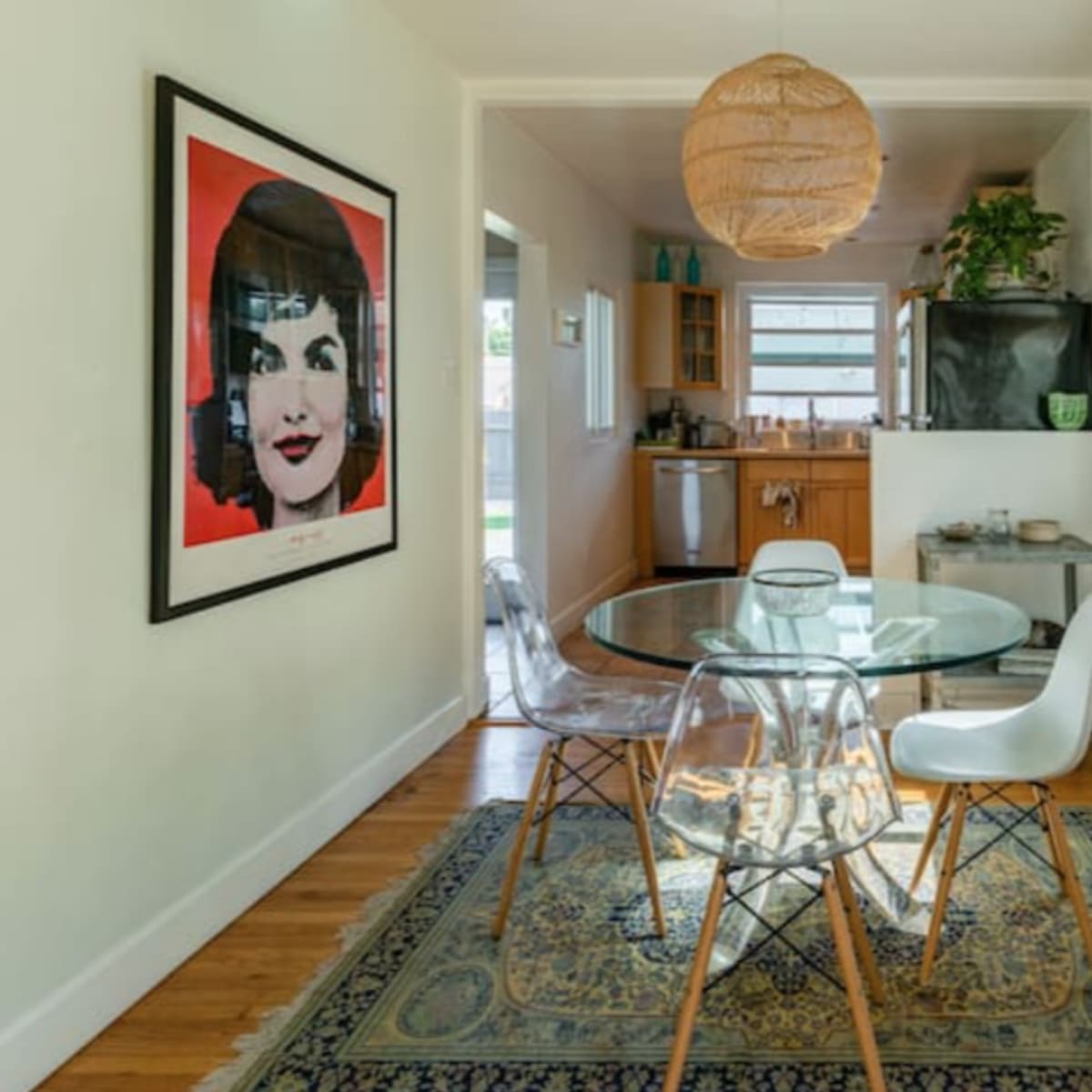 A glass dining table with four chairs is positioned in a hallway leading to a kitchen, featuring a large framed artwork and a wall display of toy cars.