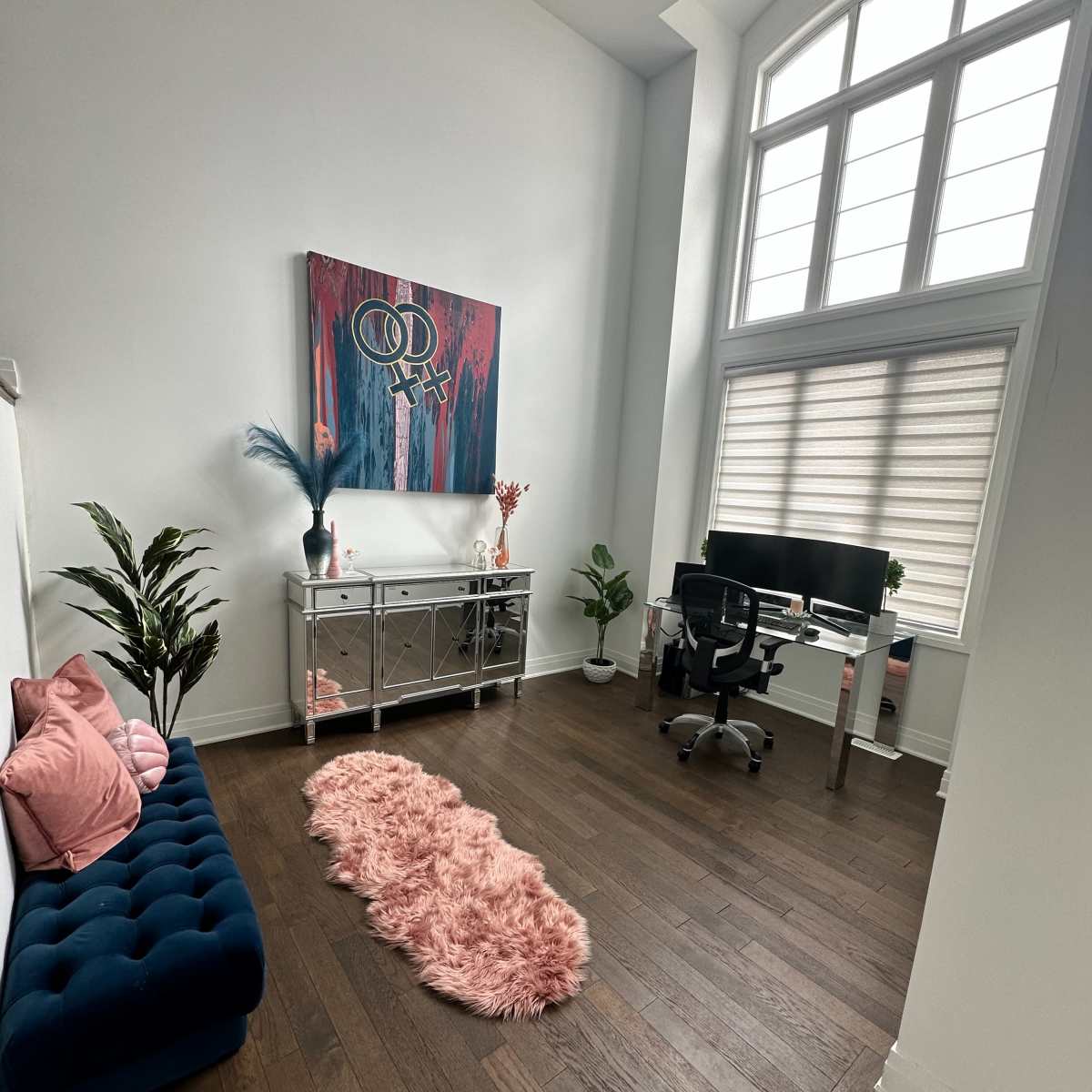 A modern office space featuring a blue tufted sofa, a glass console table, a computer desk with a black chair, a decorative rug, and two potted plants.