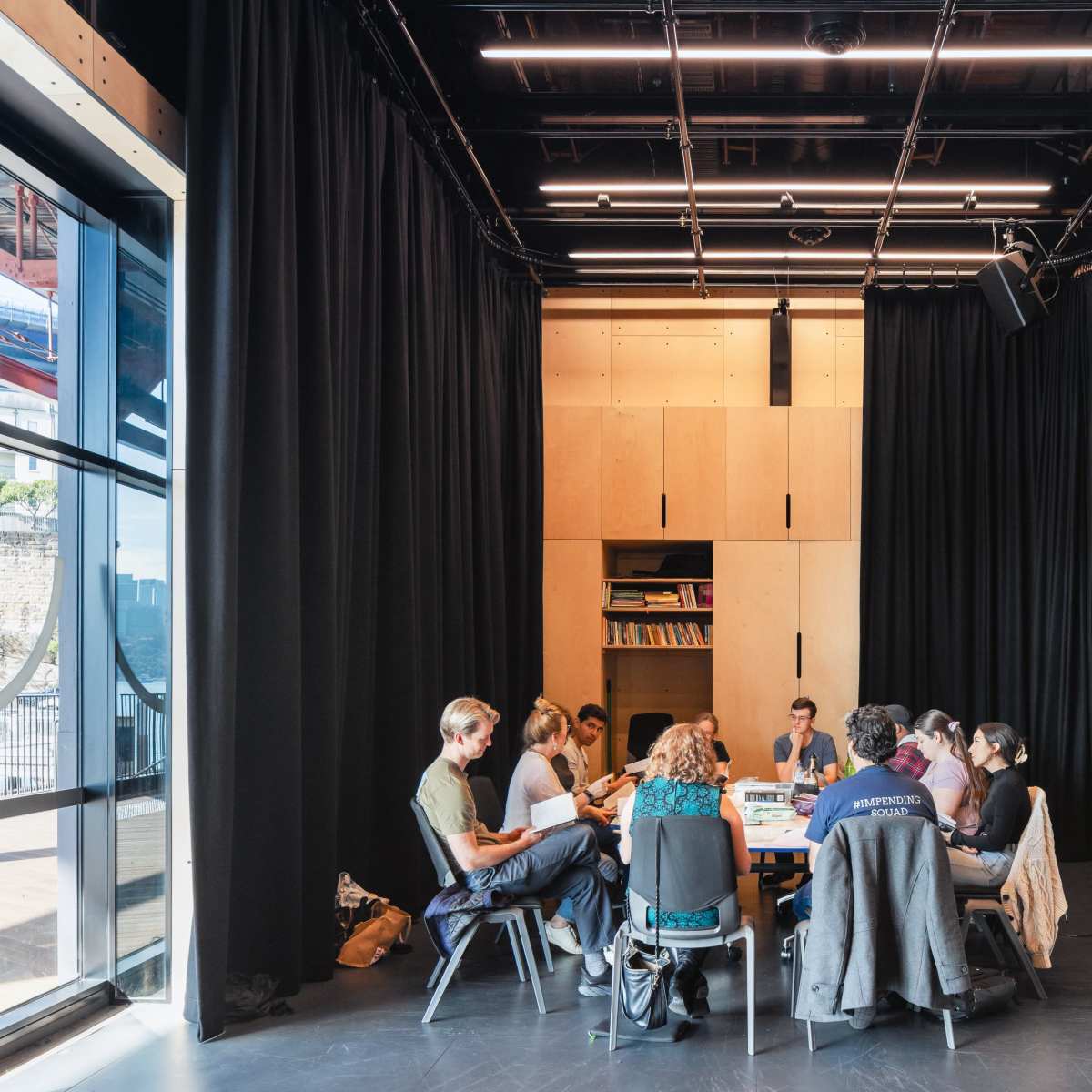 A group of people is gathered around a table in a modern, dimly-lit meeting room with large windows and black curtains.