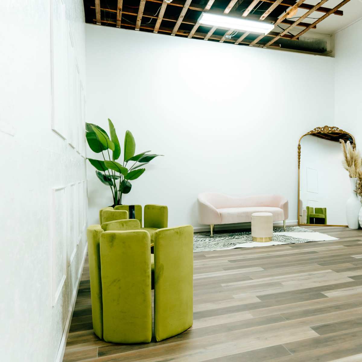 A minimalistic interior space with a pink sofa, green chairs, a decorative mirror, and indoor plants.