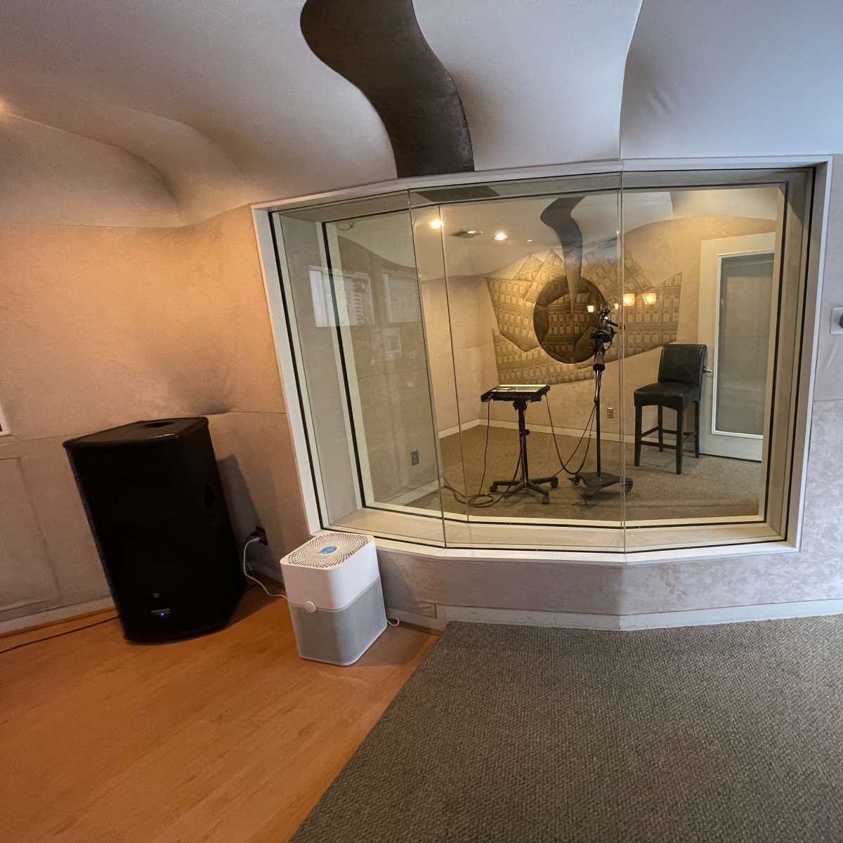 A soundproof recording booth with microphone and stand, surrounded by glass panels, located in a room with muted decor.