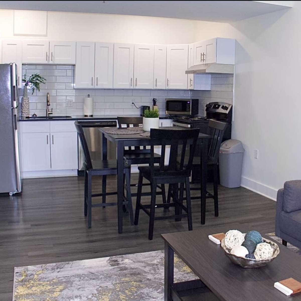 A modern kitchen and living area, featuring a dining table, black chairs, and a gray couch with decorative pillows.