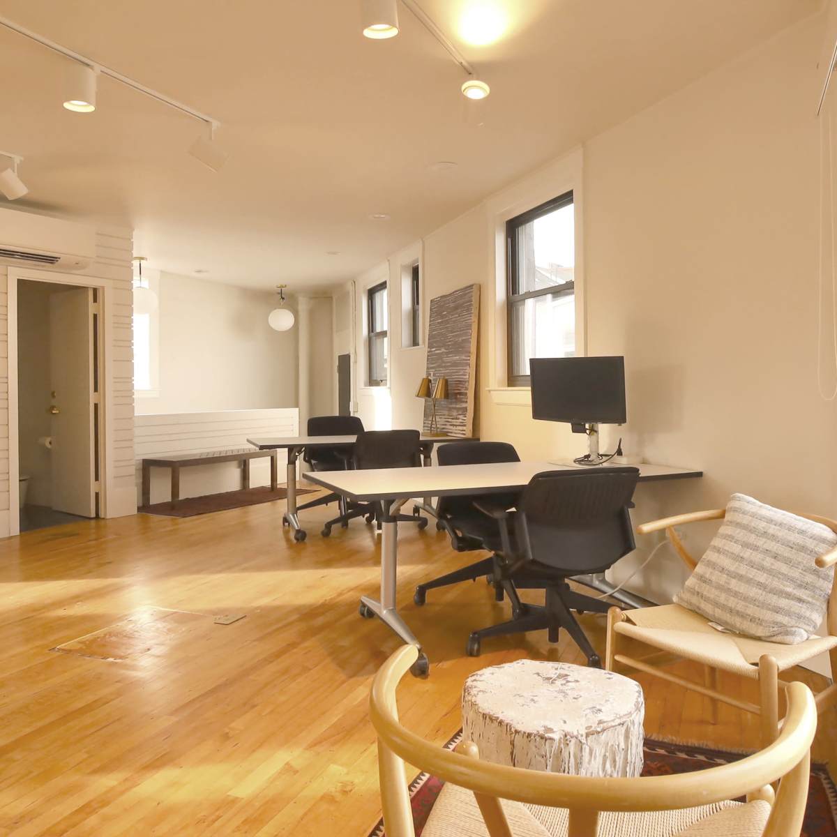 A bright, modern office space featuring wooden floors, multiple desks with chairs, and a comfortable seating area near a large window.