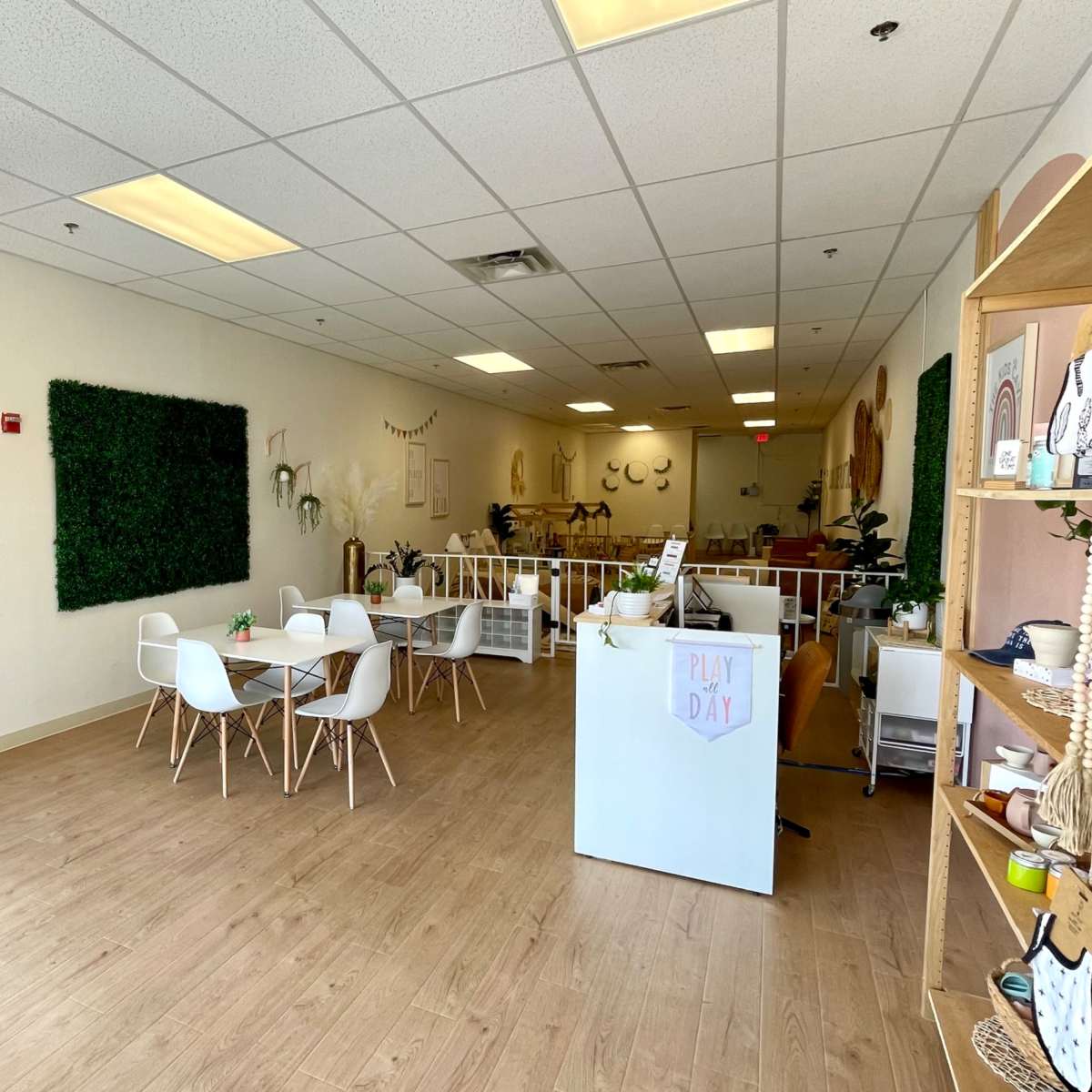 A spacious, bright interior of a room with wooden flooring, featuring a seating area with white chairs and tables, green wall decorations, and shelves displaying various items.