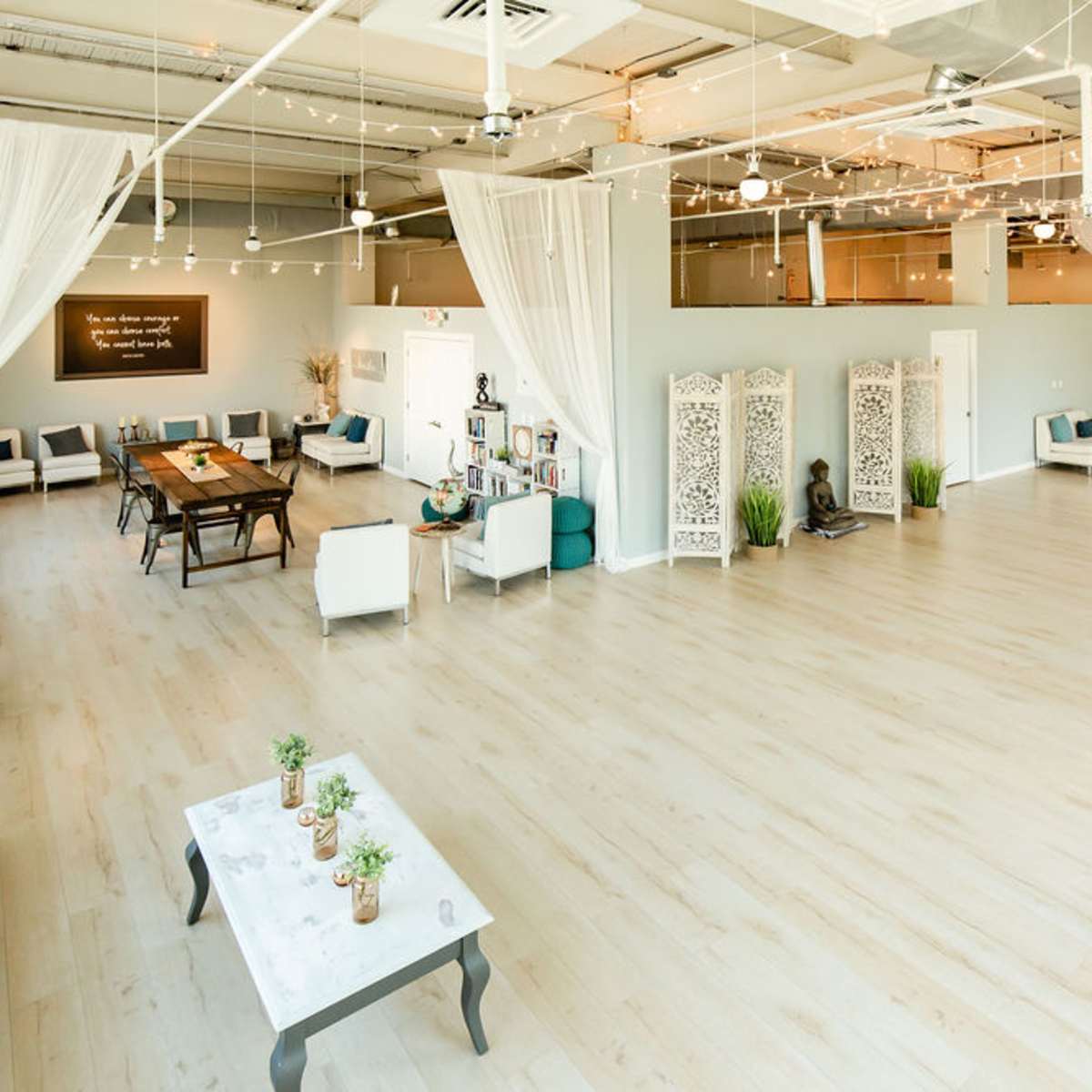A spacious, well-lit studio with wooden flooring, white drapes, and a mix of seating arrangements, including a long table and decorative screens.
