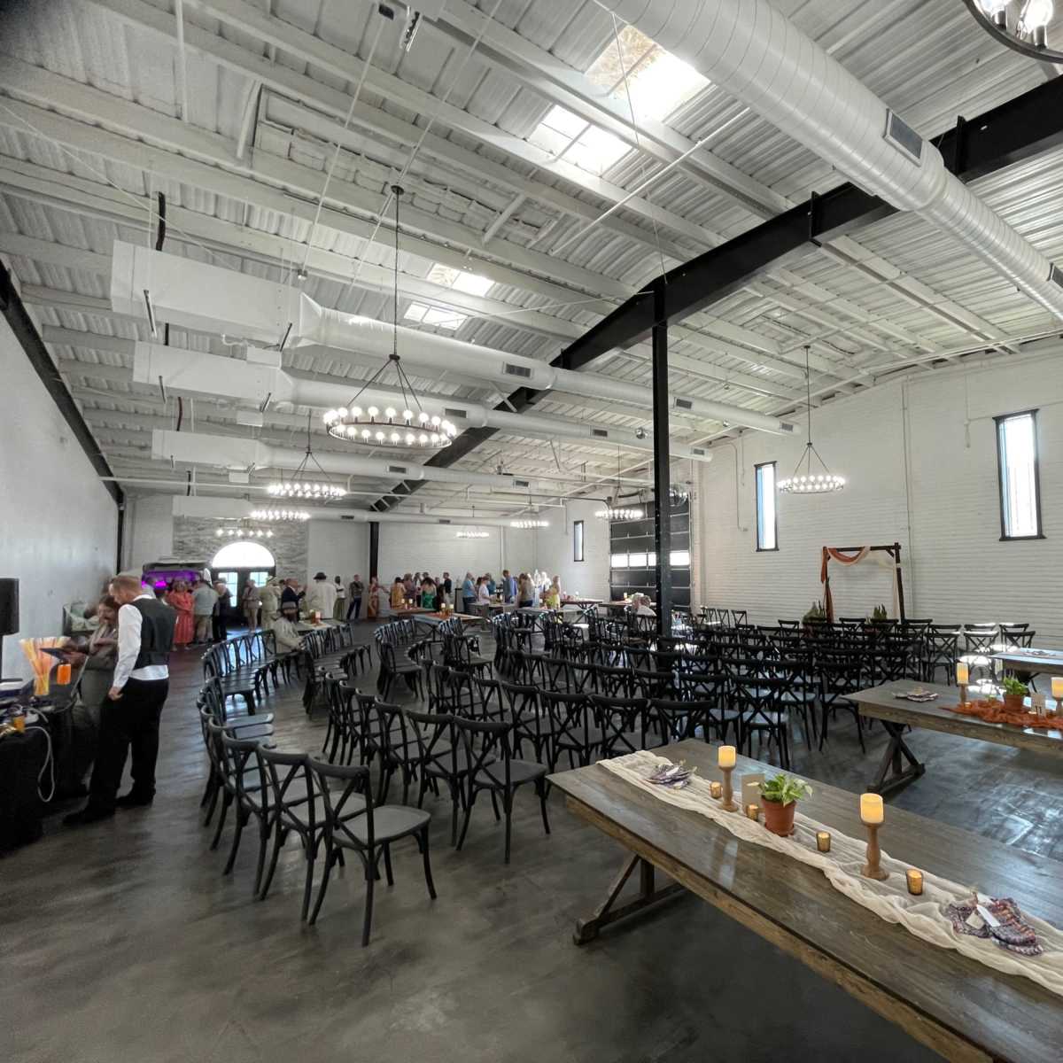 A large, industrial-style event space set up with rows of black chairs and long wooden tables, with guests mingling in the background.