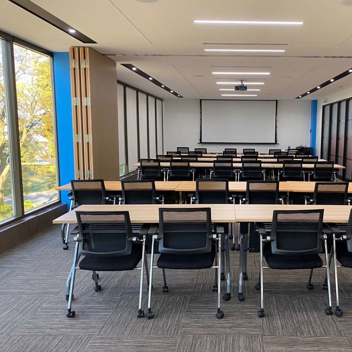A modern meeting room features rows of tables and black chairs with large windows providing natural light.