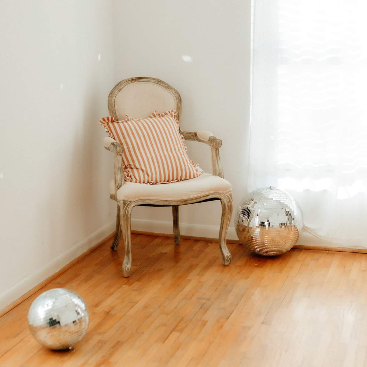 A white-walled room features a single ornate chair with a striped cushion and two disco balls on the wooden floor.