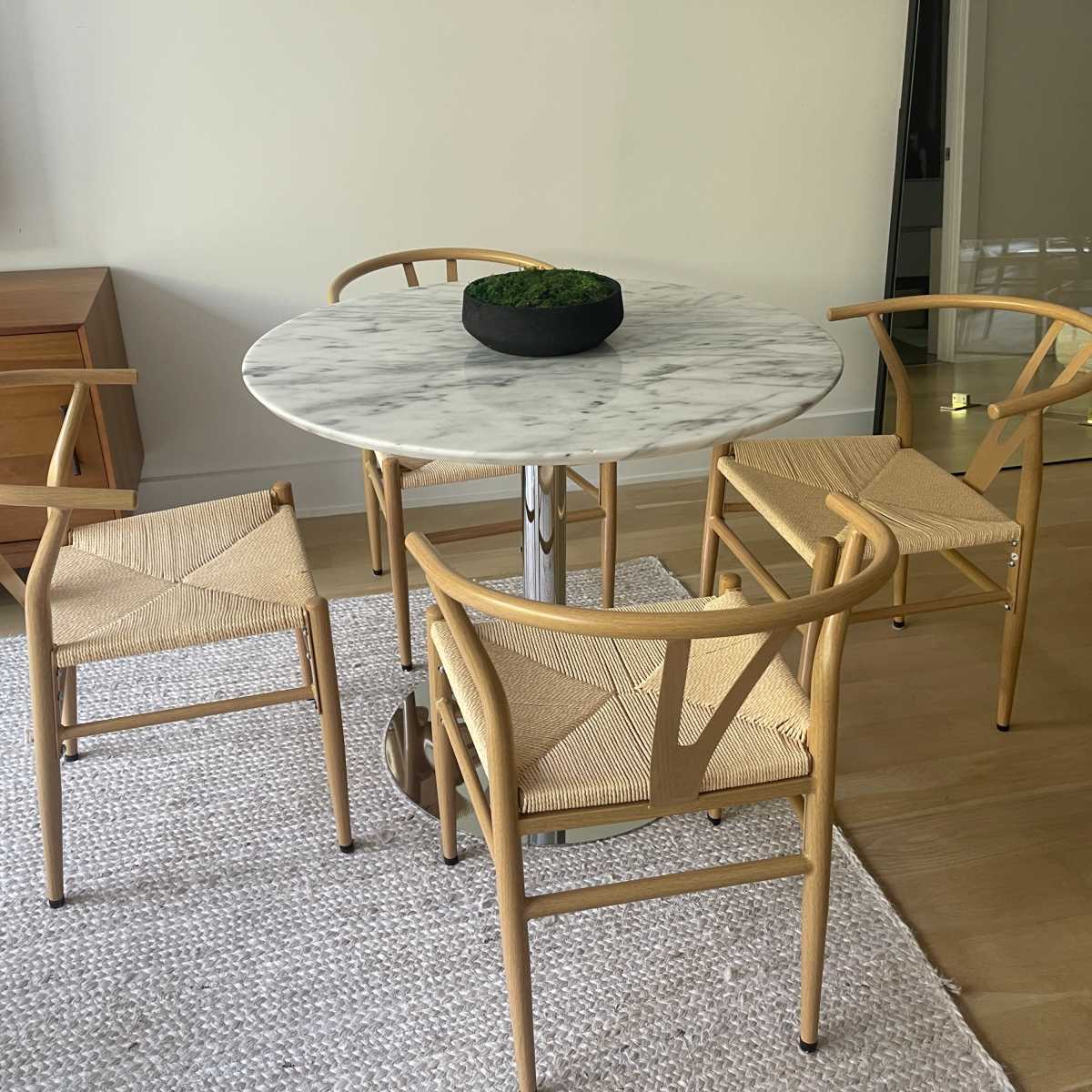 A round marble dining table is surrounded by four wooden chairs with woven seats in a minimalist interior.