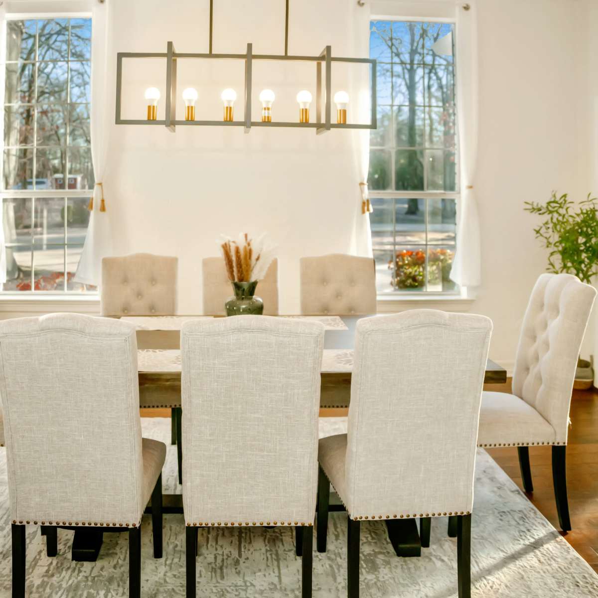 A dining room features a rectangular table surrounded by six upholstered chairs, with large windows and a hanging light fixture above.