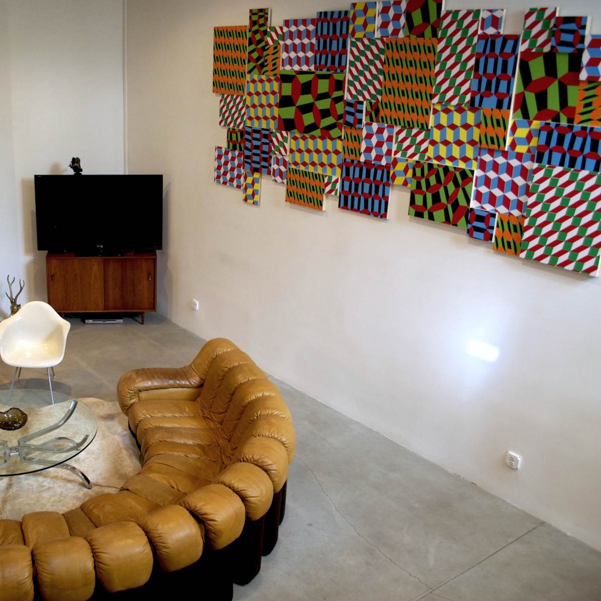 A modern living room with a curved leather sofa, a glass coffee table, a television on a cabinet, a wall of abstract art, and a geometric decoration on the ceiling.
