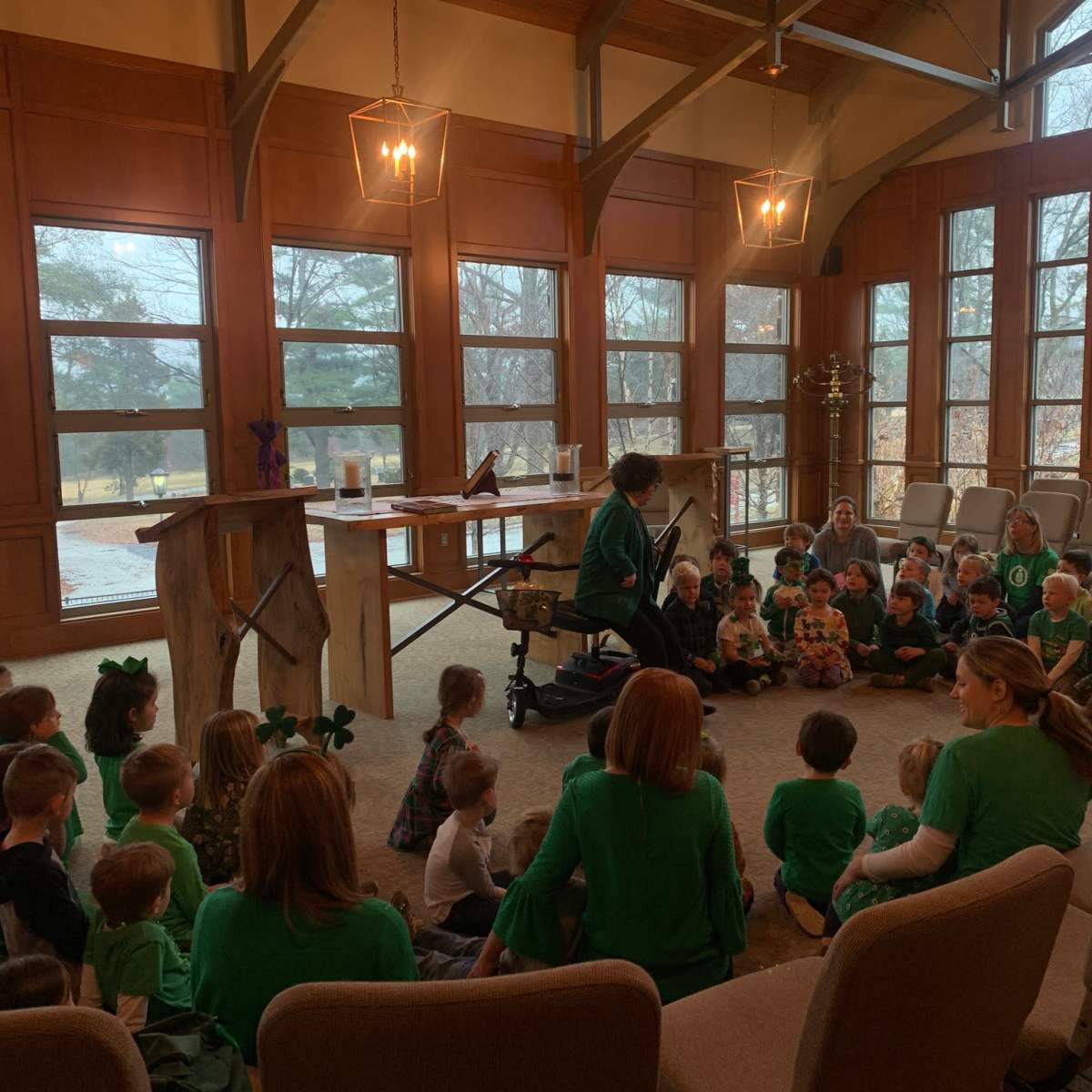 Young children in green outfits sit in a circle on the floor while an adult leads an activity in a room with large windows.