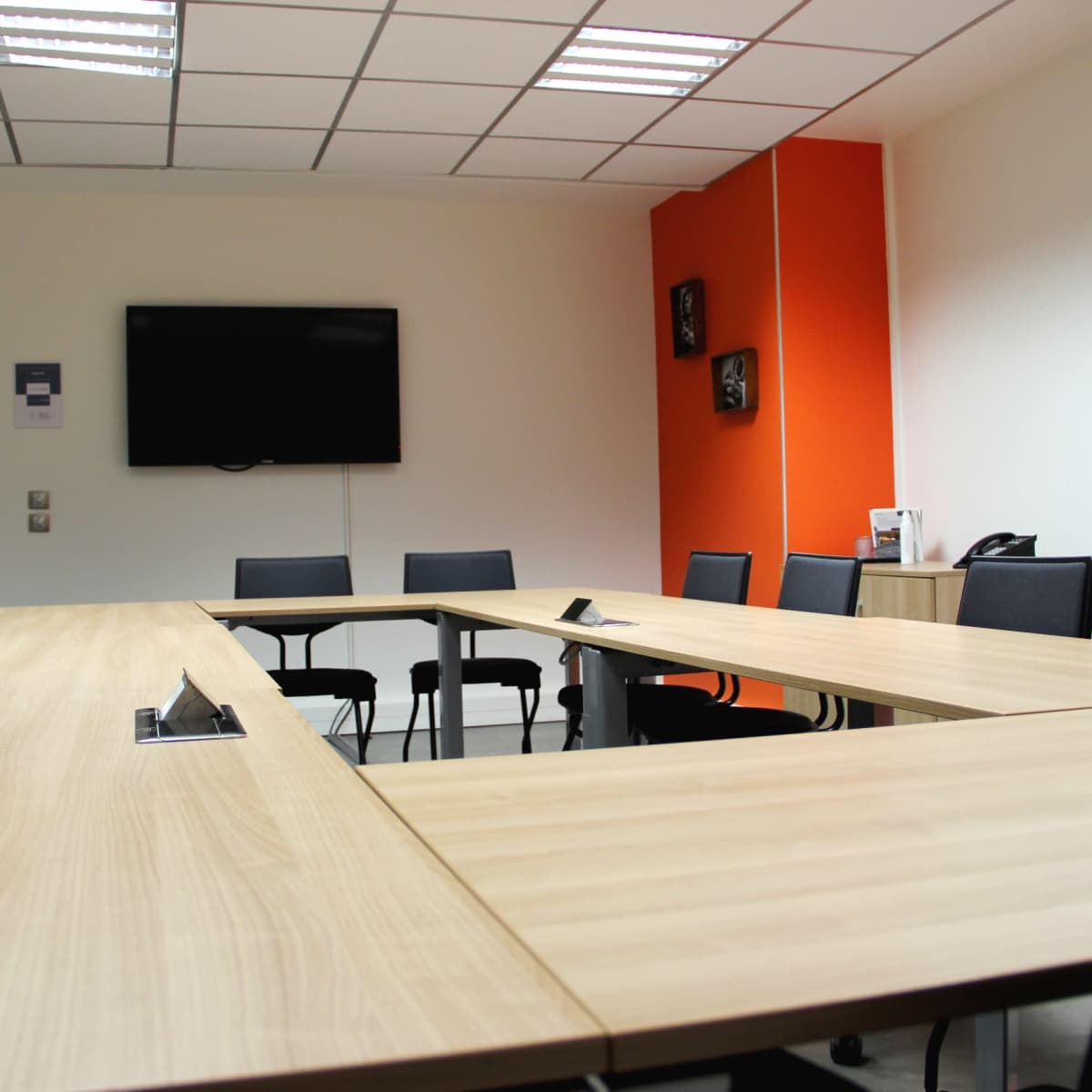 A modern conference room arranged with a large wooden table and black chairs, featuring an orange accent wall and a television mounted on one side.