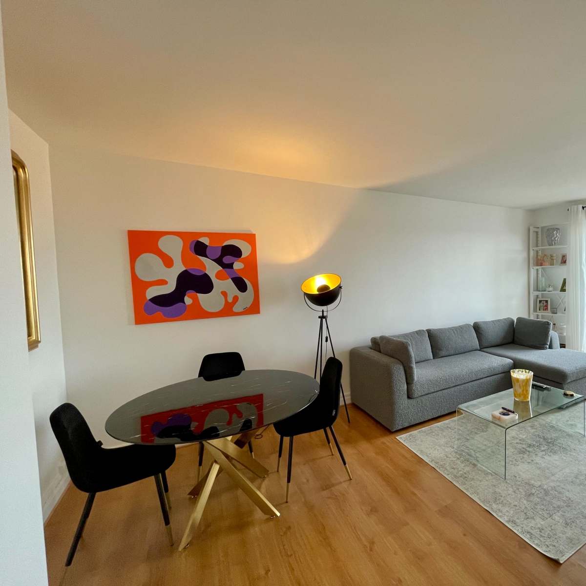 A modern living room with a gray couch, a round glass dining table, and a large window overlooking a cityscape.