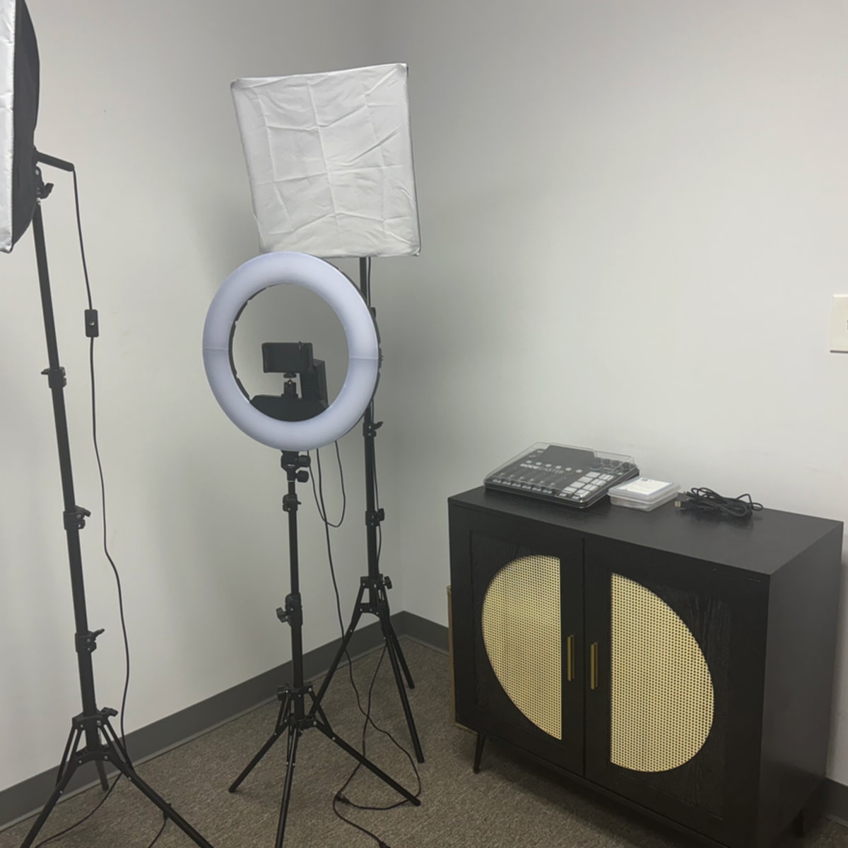 The scene shows a minimalistic setup with two softbox lights and a ring light positioned near a black cabinet and a phone on a carpeted floor in a room with white walls.