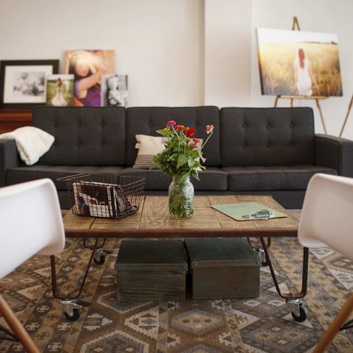 A modern living room with a gray sofa, a wooden coffee table with a vase of flowers, and two white chairs, surrounded by framed photos and artwork on easels.