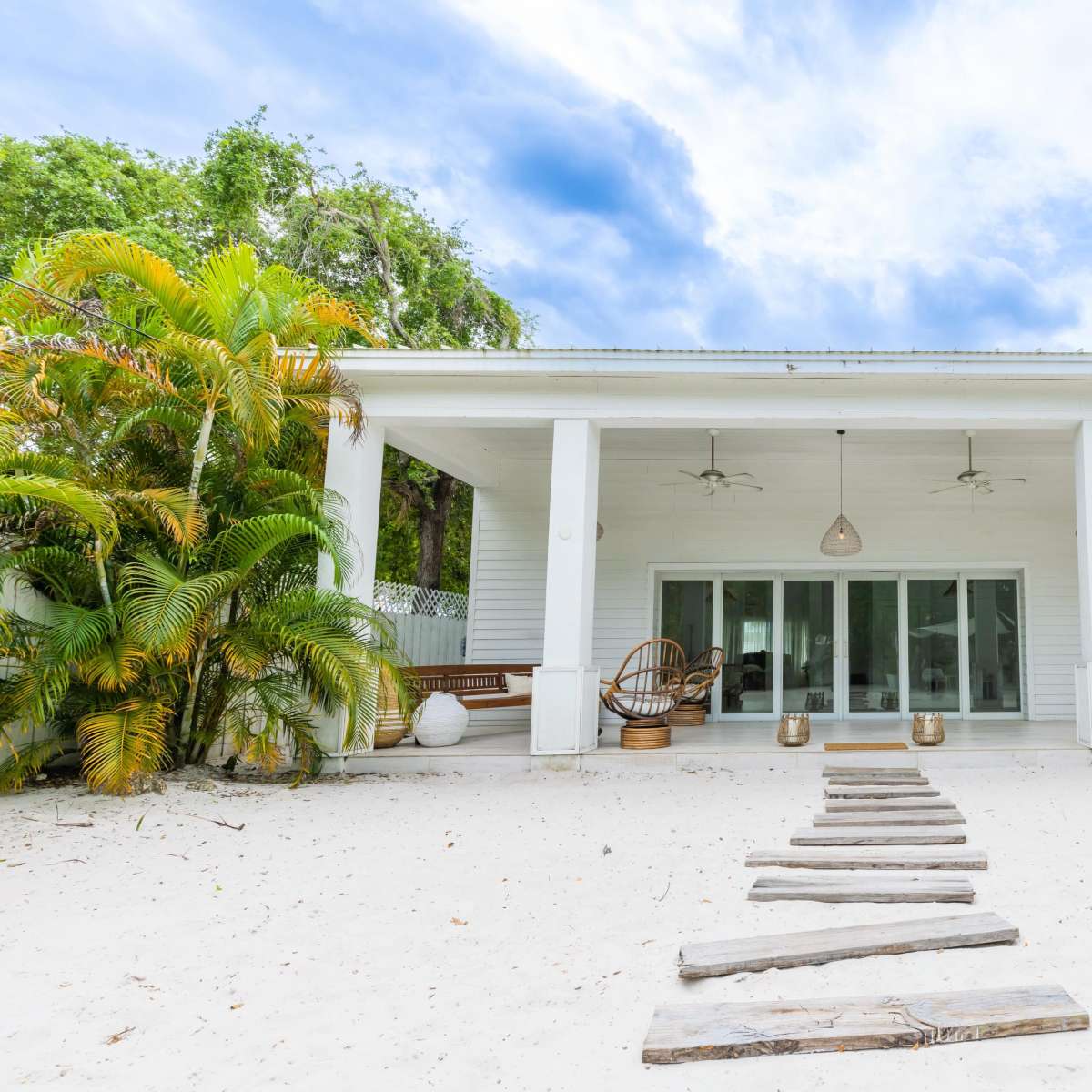 A white cottage with a porch and large glass doors sits on a sandy area surrounded by tropical greenery.