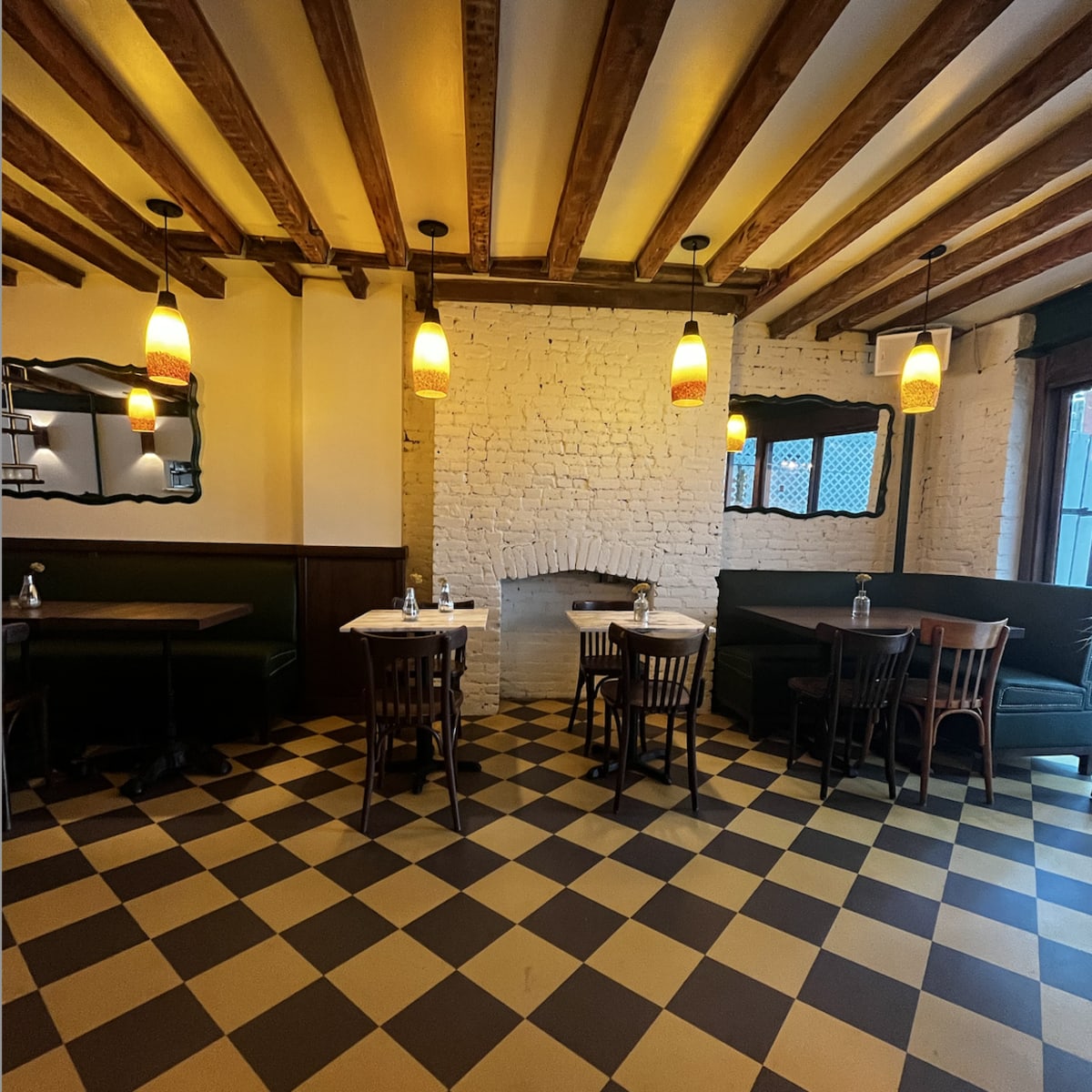 A restaurant interior with a checkerboard floor, wooden beams on the ceiling, and several tables and chairs arranged around a central fireplace.