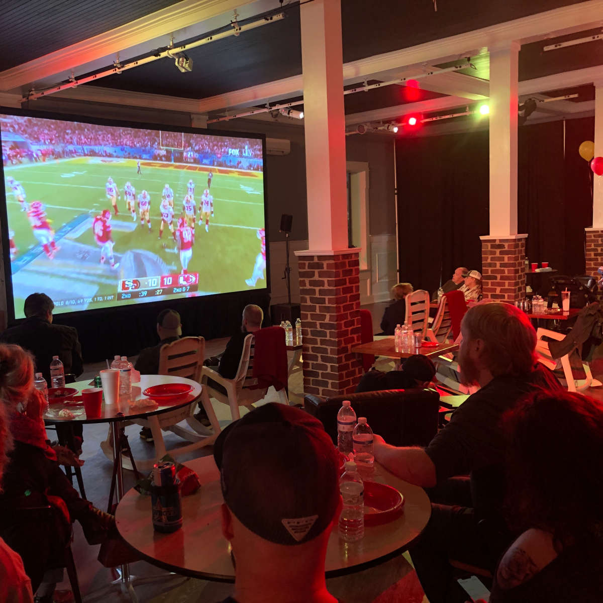 A group of people watches a football game on a large screen in a venue decorated with balloons and tables.