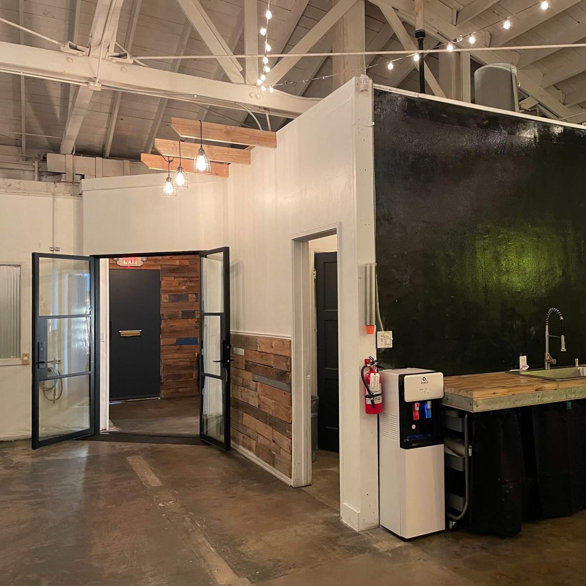 A modern interior space with a black wall, a wooden countertop with a sink, a water dispenser, and a doorway leading to another room.