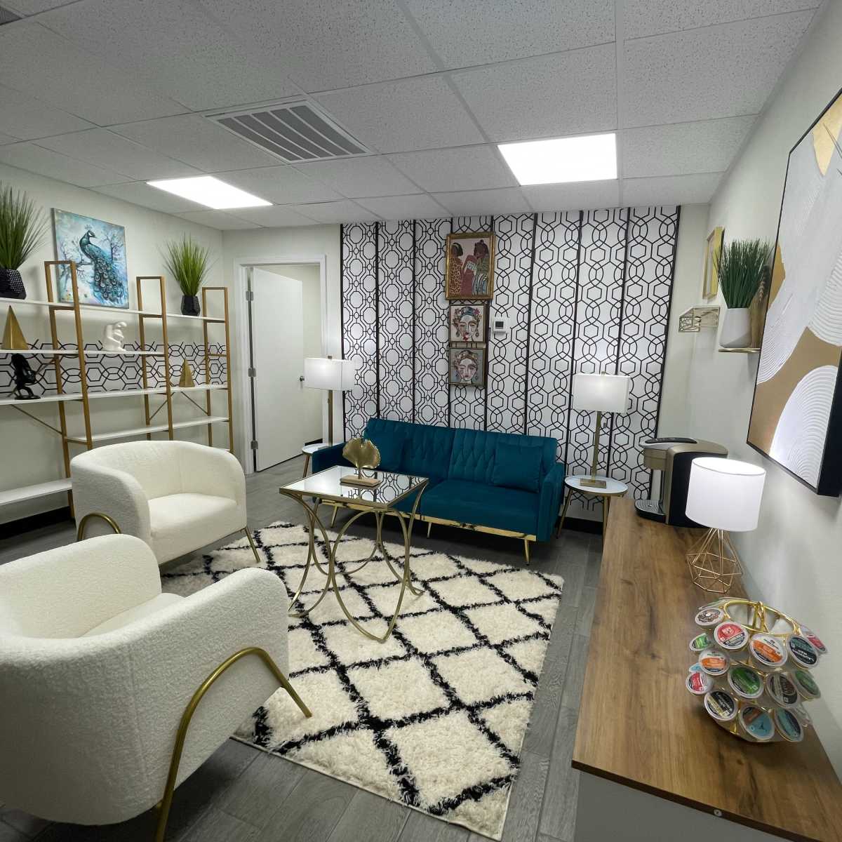 A modern lounge area with a teal sofa, two white armchairs, a coffee table, and decorative plants on shelves against a patterned wall.