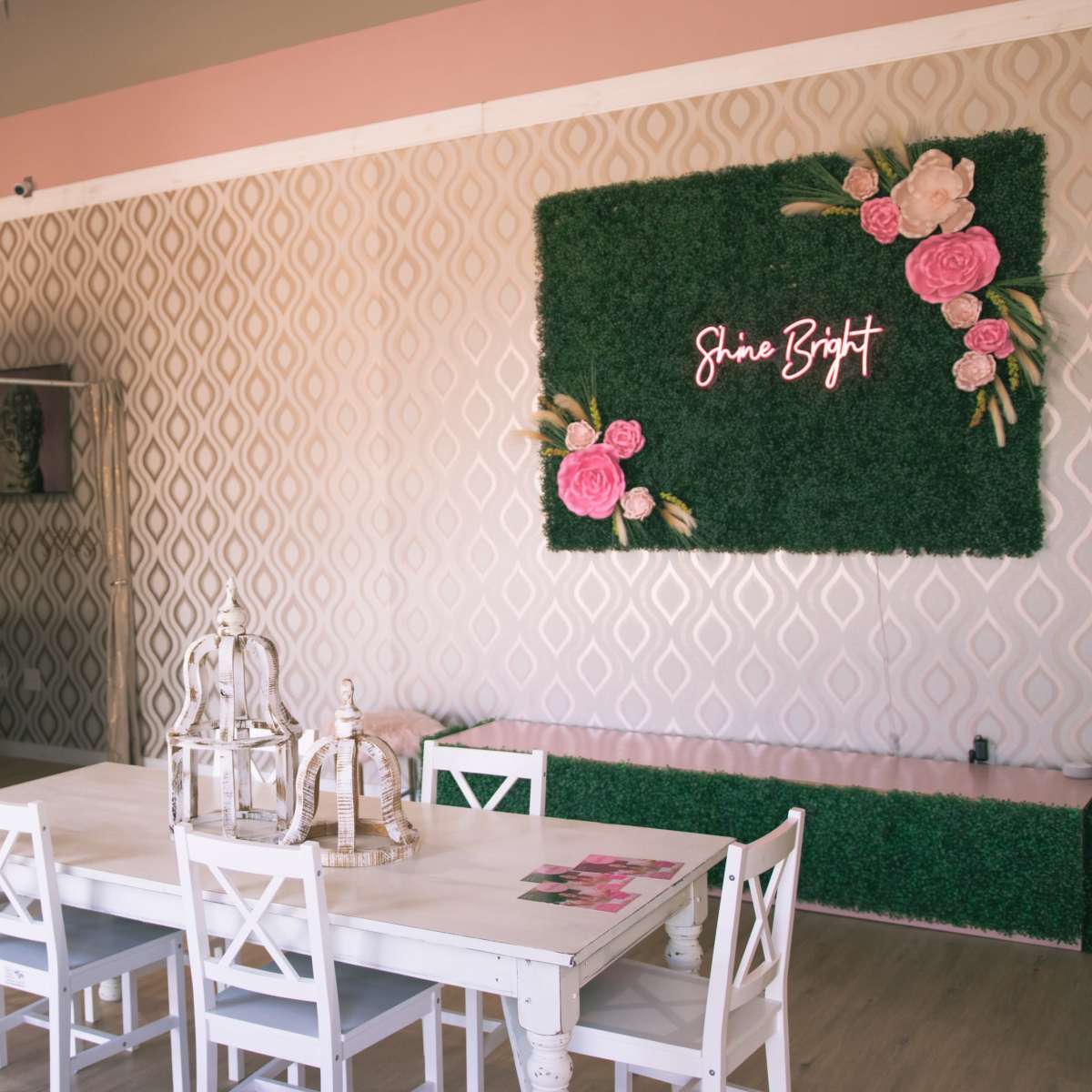 A softly decorated indoor space featuring a table with white chairs, a green wall with pink flowers, and a neon sign that reads "Shine Bright."