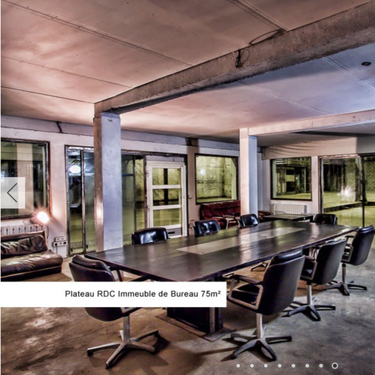 A modern office space with a large rectangular table surrounded by black chairs, concrete walls, and large windows.