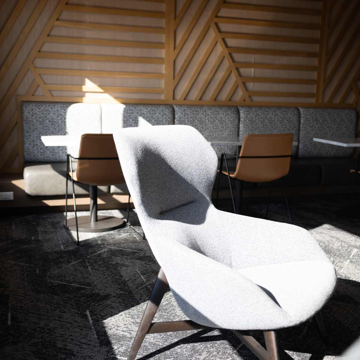 A gray lounge chair is positioned in a well-lit corner of a modern seating area with textured wooden walls and a row of tables and chairs in the background.