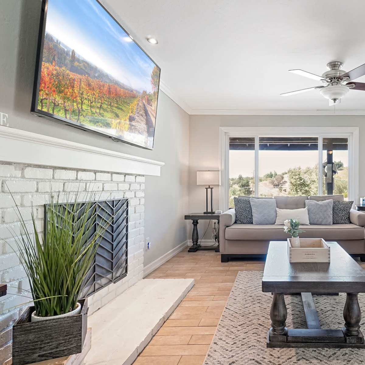 A modern living room with a sectional sofa, a coffee table, and a television mounted above a white brick fireplace, all set against neutral-colored walls and trimmed windows.