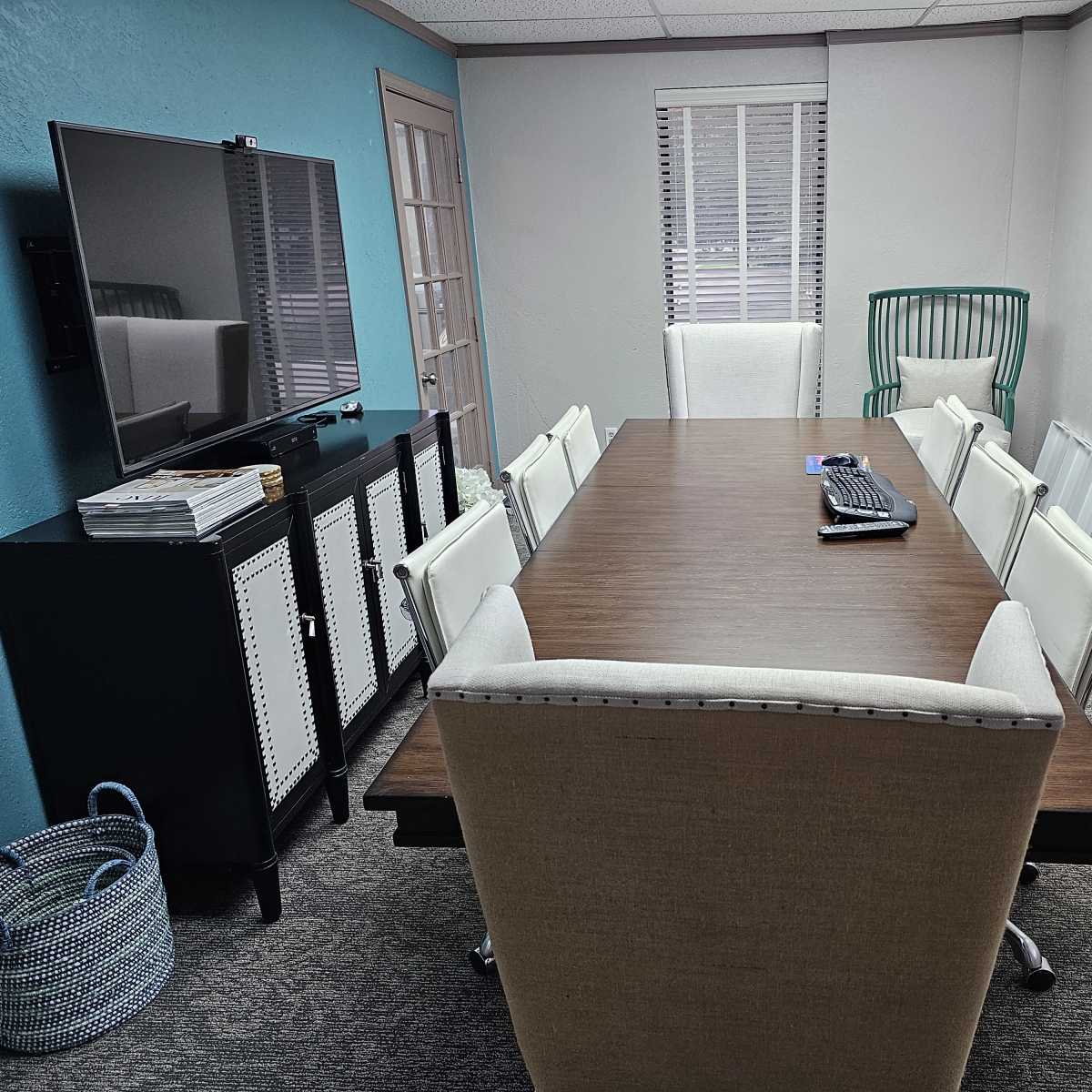 A modern conference room with a long wooden table surrounded by white chairs, a large television mounted on the wall, and a green accent chair in the corner.