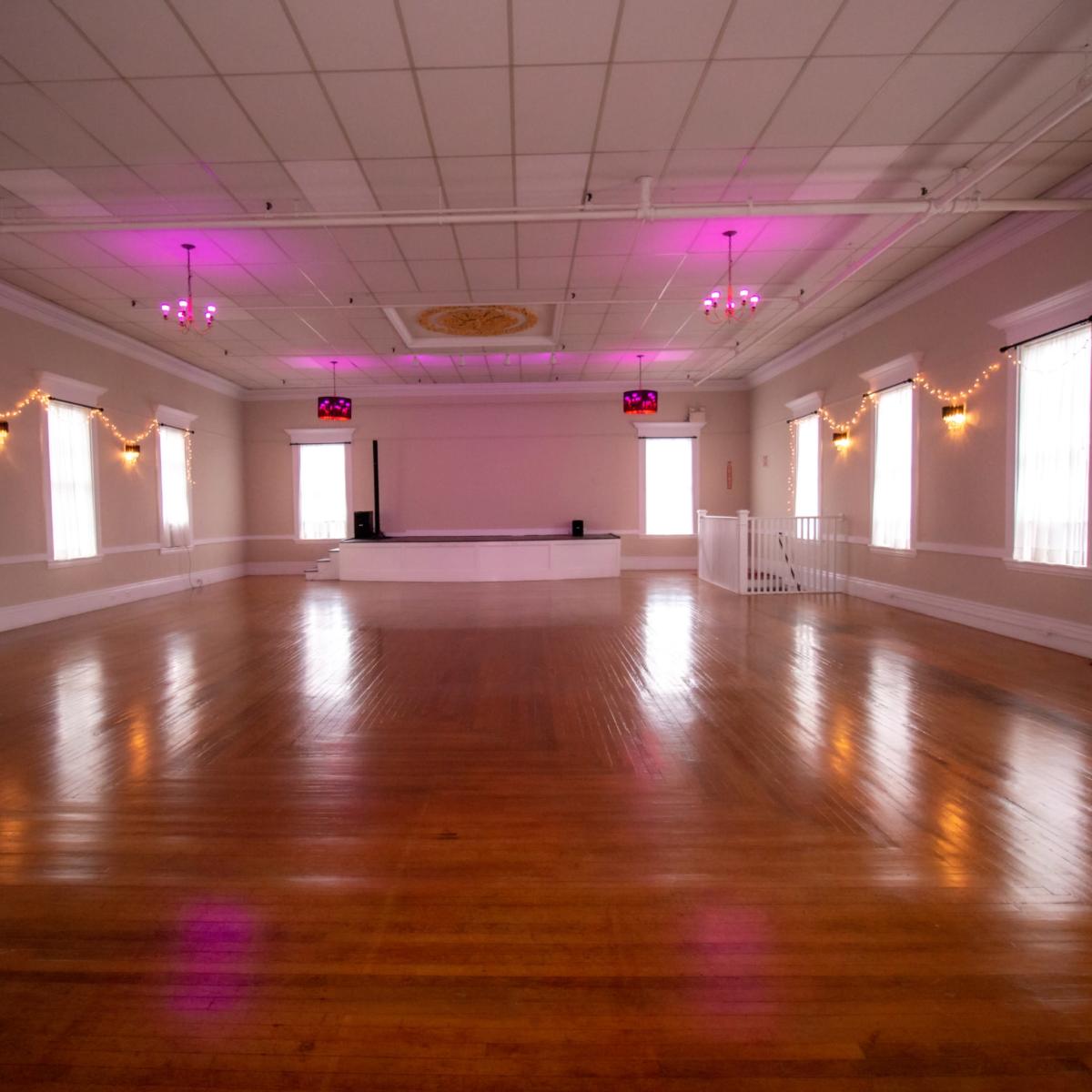A spacious, well-lit event hall with a polished wooden floor, accented by pink lighting and decorative string lights along the walls.