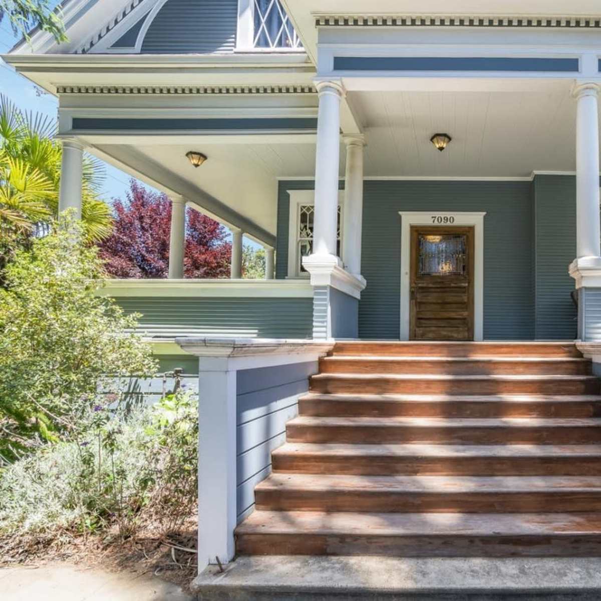 A set of wooden steps leads up to a blue house with a front door framed by white columns and greenery on either side.