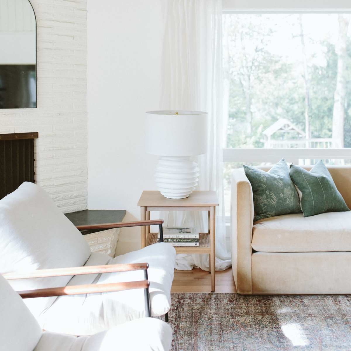 A cozy living room features a light-colored sofa, a decorative lamp on a side table, and large windows with sheer curtains allowing natural light to flow in.