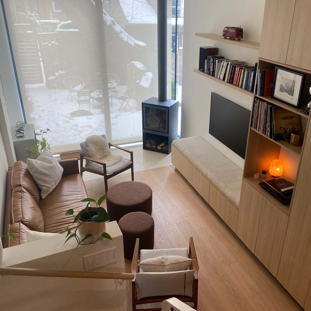 A cozy living space featuring a brown leather sofa, a wooden bookshelf with decor, a television, and a view of a winter scene through a large window.