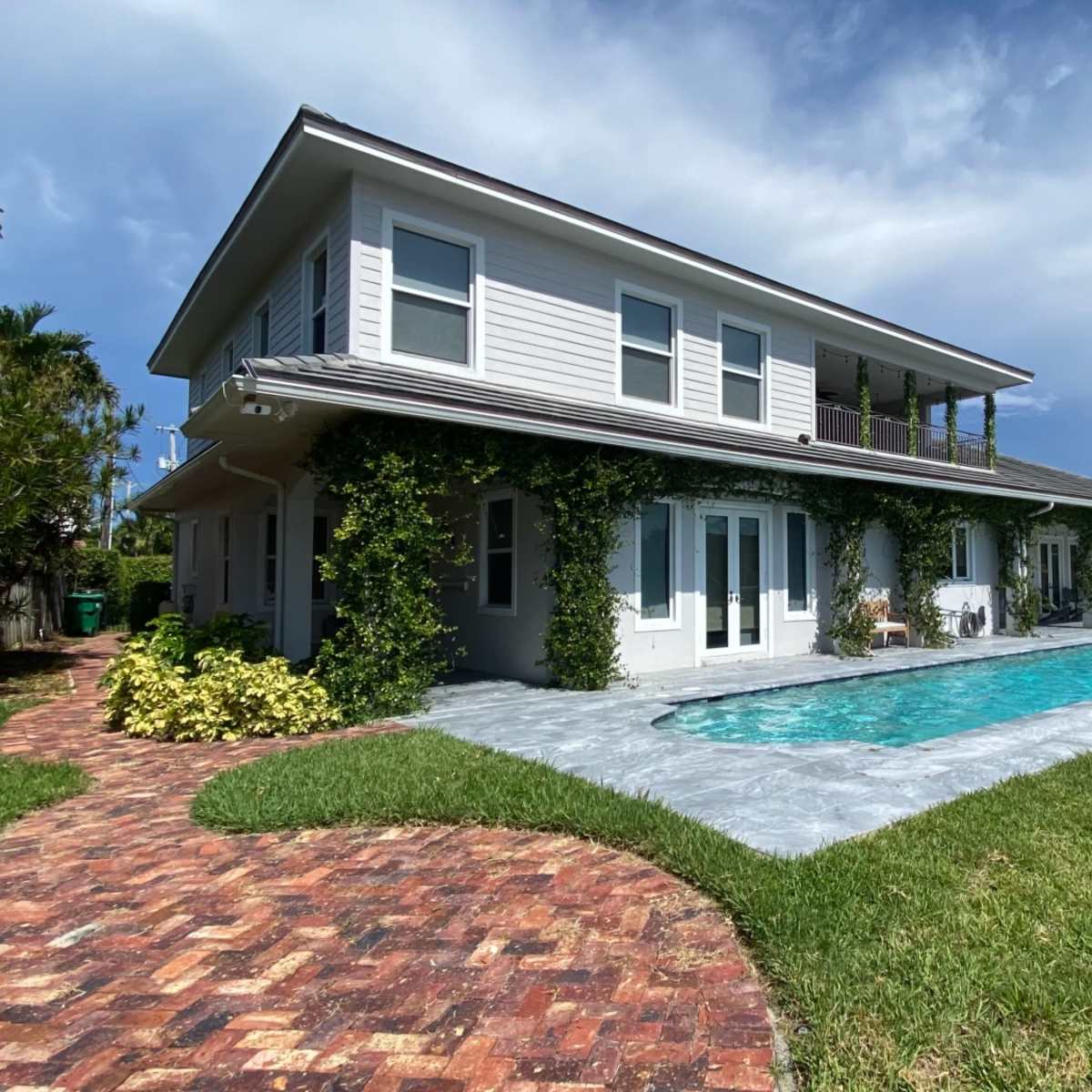 A two-story house with a pool and landscaped gardens is outlined by brick pathways and surrounded by palm trees.