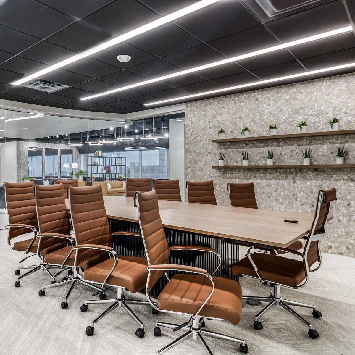 A modern conference room features a long wooden table surrounded by brown leather chairs, with a glass wall and a plant in the corner.