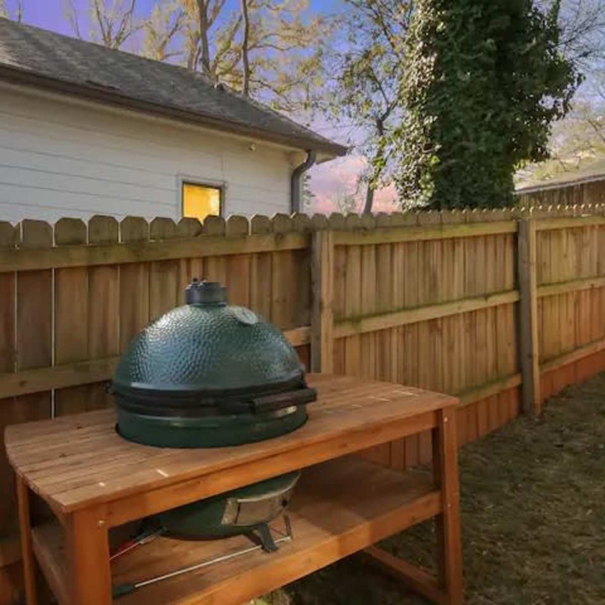 A green ceramic grill sits on a wooden table next to a wooden fence in a backyard at dusk.