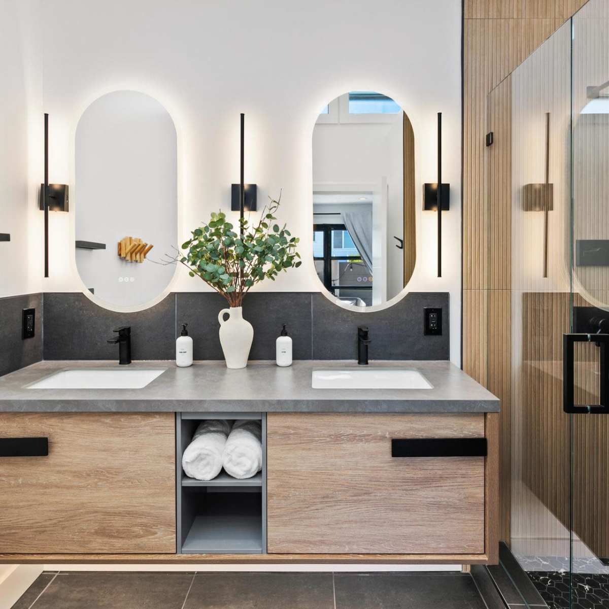 A modern bathroom featuring a dual-sink vanity with a concrete countertop, wooden cabinets, and two round mirrors illuminated by wall sconces, alongside a glass shower enclosure.