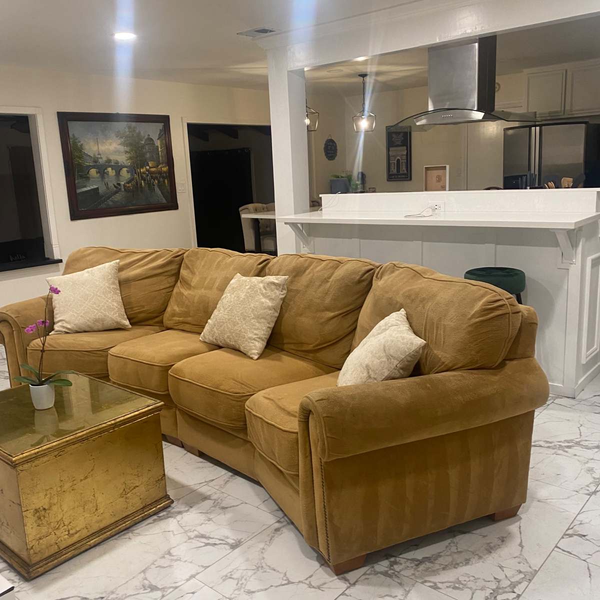 A tan sectional sofa with decorative pillows in a living area featuring marble-like flooring and a kitchen with a white bar counter in the background.