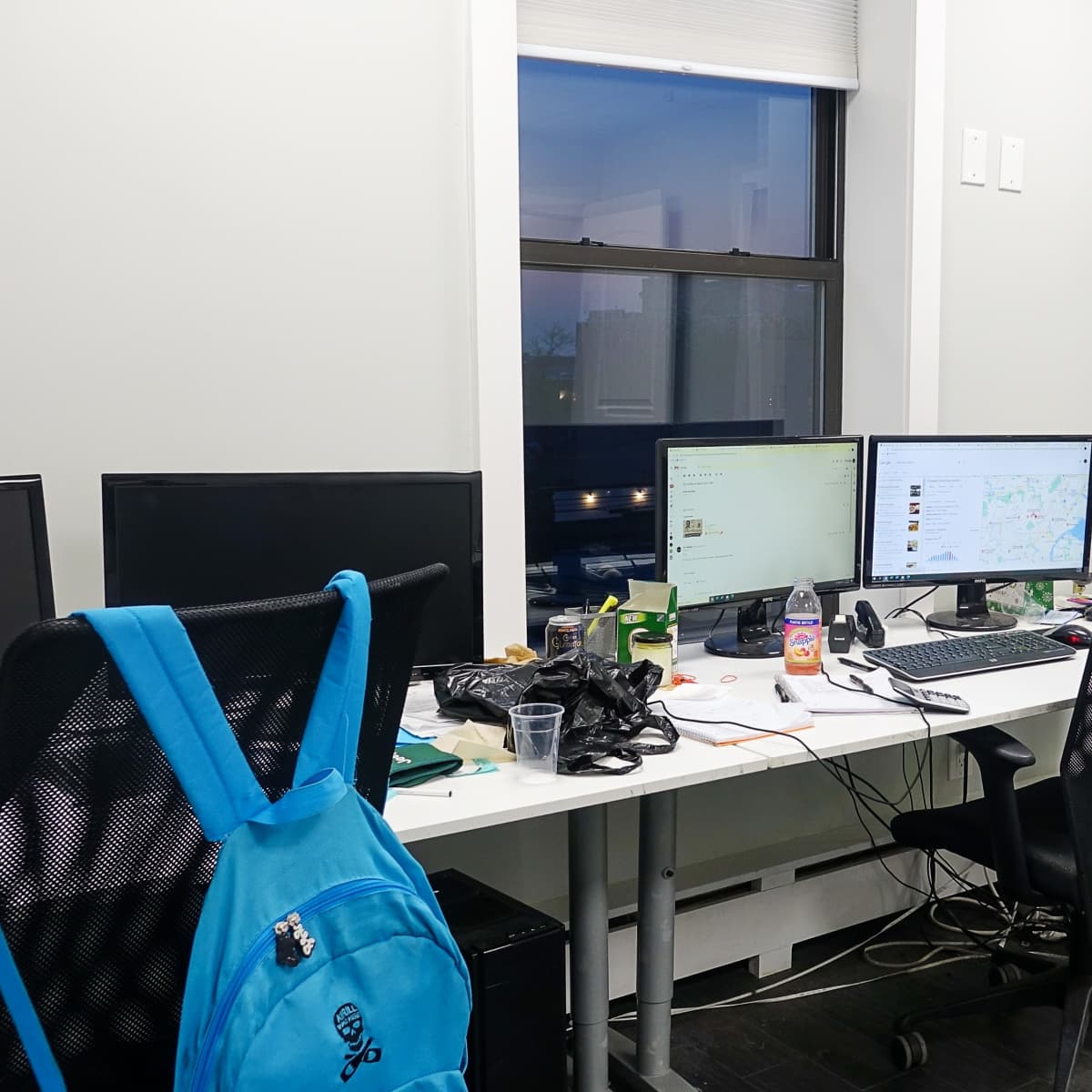 An office space with multiple computer monitors, a printer, and a desk cluttered with various items, including a backpack and drinks.