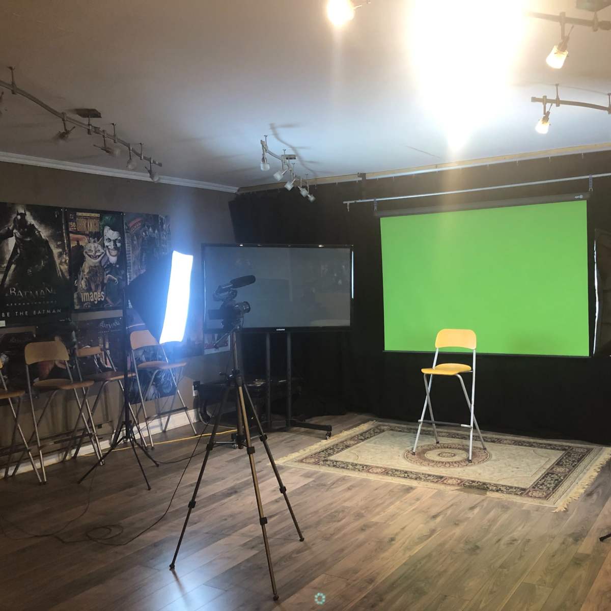 A video recording studio is set up with a green screen, a chair, lighting equipment, and cameras, all arranged in a spacious room with dark curtains and movie posters on the walls.