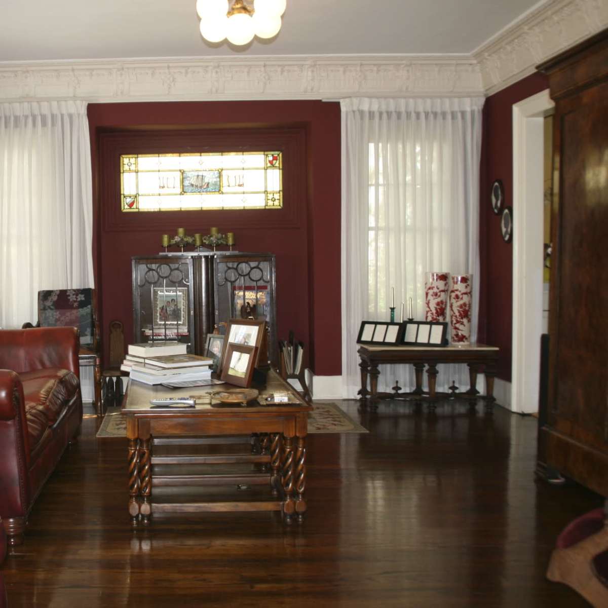 A living room with a red leather sofa, a wooden coffee table, large windows dressed with sheer curtains, and decorative cabinets against a dark red wall.